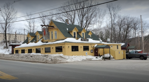 A yellow, two-story building with a green roof, surrounded by snow, located on a roadside in a winter setting.