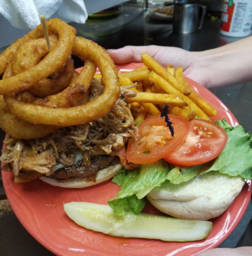 A hand holds a plate with a stacked burger topped with onion rings, pulled pork, lettuce, tomato, and fries on the side.