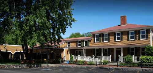 A large yellow house with a red roof, surrounded by trees and a parking area, featuring a porch and white railings.