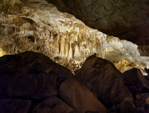 Stalactites hang from a cave ceiling, illuminated by soft lighting, with large rocks in the foreground.