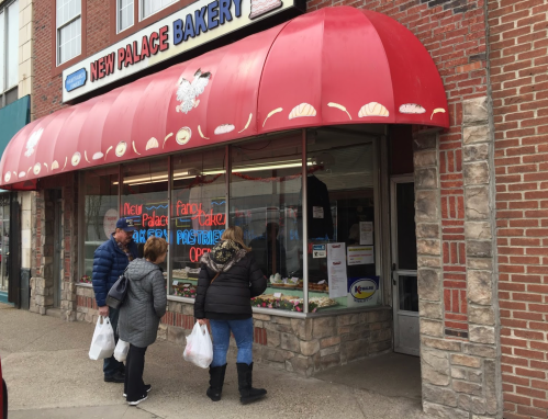 A bakery storefront with a red awning, featuring customers looking at pastries through the window.