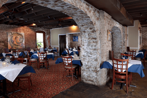 Cozy restaurant interior with stone arches, wooden tables, and blue tablecloths, featuring artwork on the walls.