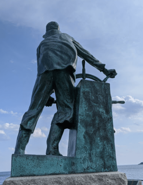 A bronze statue of a sailor at the helm, facing the sea under a blue sky with scattered clouds.
