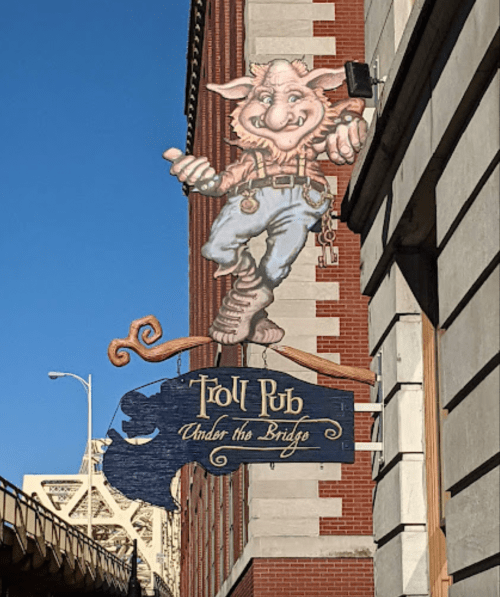 A whimsical sign featuring a cartoon troll in overalls, advertising "Troll Pub Under the Bridge" on a brick building.