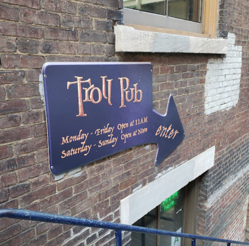 Sign for Troll Pub with opening hours and an arrow pointing to the entrance, mounted on a brick wall.