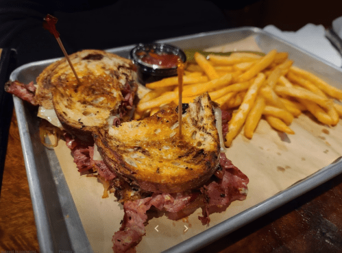 A grilled sandwich with layers of meat and cheese, served with fries and a small bowl of salsa on a metal tray.