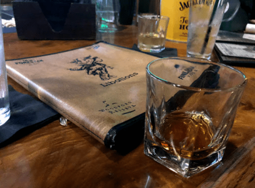 A whiskey glass sits next to a menu on a wooden table, with drinks and glasses in the background.