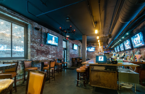 Interior of a bar with exposed brick walls, wooden stools, and multiple TVs displaying sports. Warm lighting creates a cozy atmosphere.
