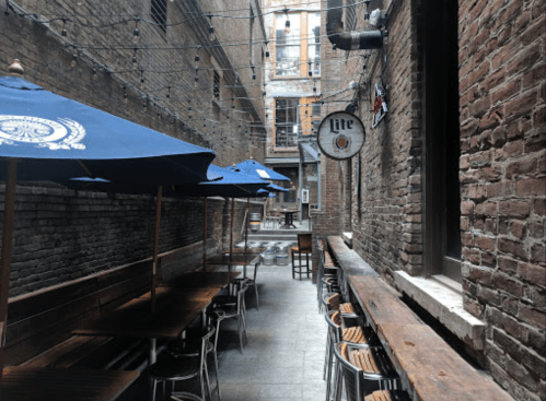 Narrow alleyway with wooden tables, blue umbrellas, and brick walls, creating a cozy outdoor dining space.