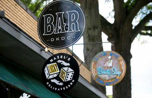 Signage for a bar and a delicatessen, with a beauty shop sign in the background, under trees.