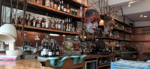 A bar with shelves filled with various bottles, decorative masks, and a wooden counter with a cash register.