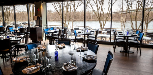 A modern restaurant interior with round tables set for dining, large windows overlooking a river and trees outside.