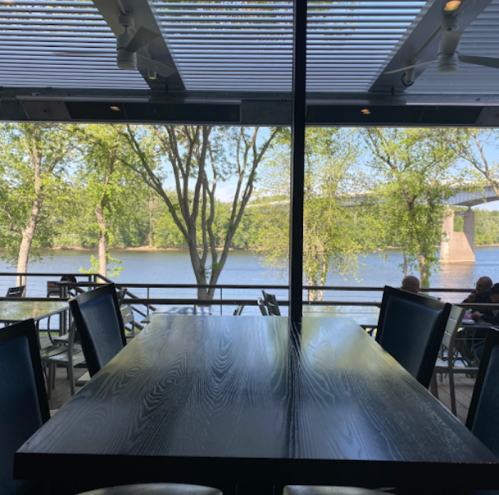 A view from inside a restaurant, featuring a wooden table and large windows overlooking a river and trees.