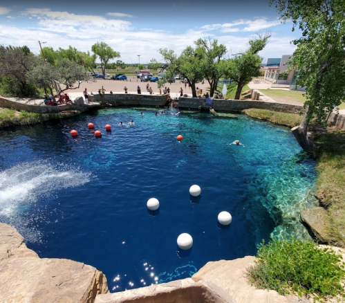 A clear blue swimming hole with people swimming, surrounded by trees and a grassy area, and buoy markers in the water.