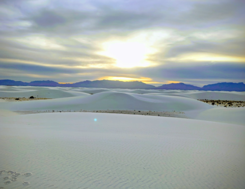 A serene desert landscape with white sand dunes under a cloudy sky, illuminated by a soft sunset in the background.