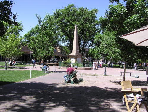 A park scene with an obelisk monument surrounded by trees, benches, and people enjoying the outdoors on a sunny day.