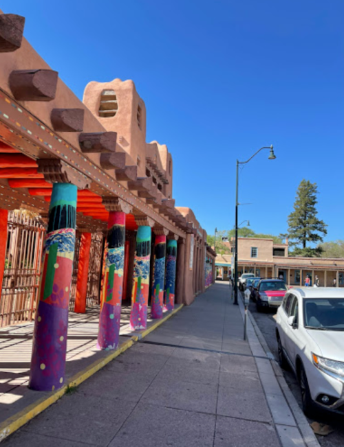 Colorful, painted columns line a sidewalk next to a building with a red roof under a clear blue sky.