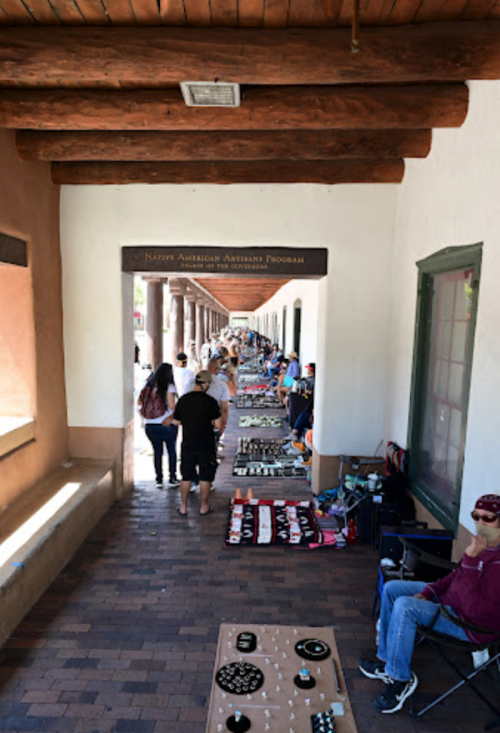 A bustling market scene with vendors displaying crafts along a corridor, people browsing and shopping.