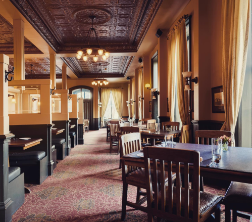 Elegant restaurant interior with wooden tables, chairs, and ornate ceiling, featuring warm lighting and large windows.