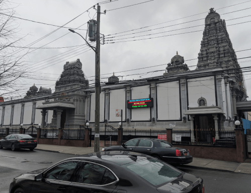 A large temple with intricate architecture, surrounded by cars and power lines, under a cloudy sky.