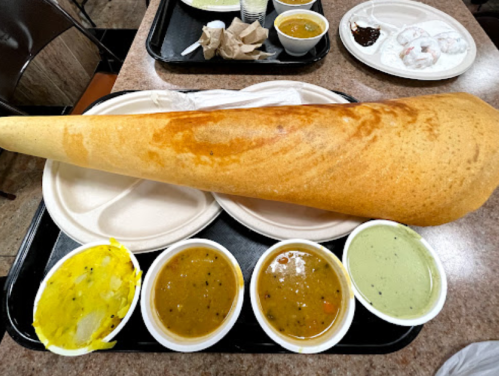 A large dosa on a tray with four small bowls of various chutneys and sambar.