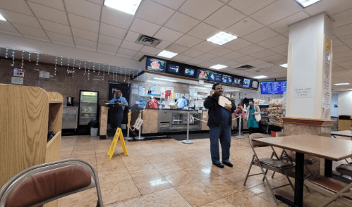 A cafeteria with a counter, menu screens, and two staff members; a caution sign is visible on the floor.