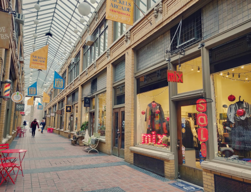 A charming arcade with shops, colorful signs, and red chairs lining the walkway, under a glass roof.