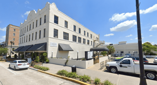 A three-story building with a white facade, featuring awnings and parked cars in front under a blue sky.