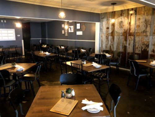A cozy restaurant interior with wooden tables, black chairs, and warm lighting, featuring a rustic wall design.
