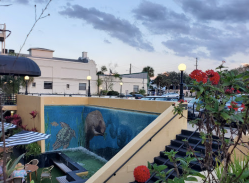 A mural of an elephant in a pool area, surrounded by plants and a staircase, with a cloudy sky above.