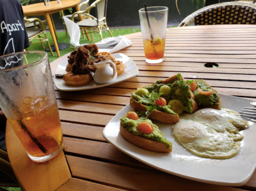 A wooden table with two plates of food: one with avocado toast and eggs, the other with fried chicken and waffles. Drinks are nearby.
