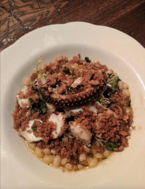 A plate of octopus atop a bed of beans and greens, garnished with crispy breadcrumbs.