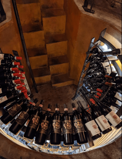 A spiral wine rack filled with various bottles, leading to a staircase in a dimly lit cellar.