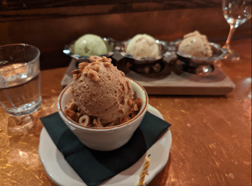 A bowl of chocolate ice cream topped with nuts, with three additional ice cream scoops in the background.