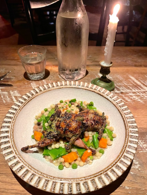 A plate of roasted quail on a bed of vegetables and couscous, with a candle and water bottle in the background.