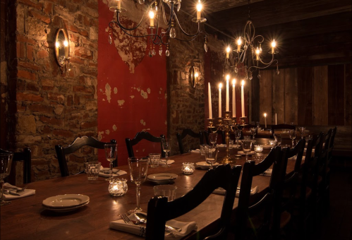 A dimly lit dining room with a long wooden table, elegant place settings, and candles on a rustic stone wall backdrop.