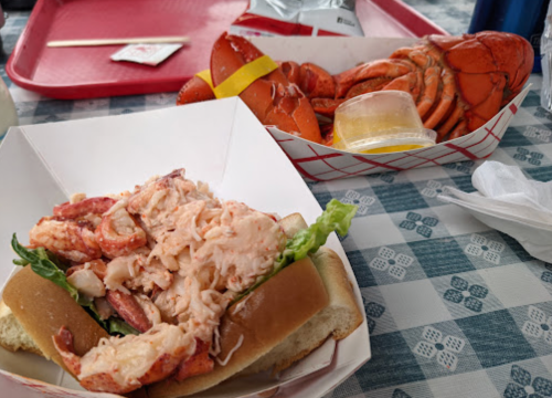 A lobster roll with lettuce on a bun, next to a tray of whole lobsters and a small container of butter.