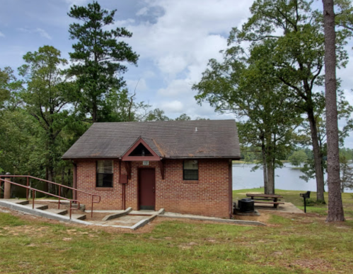 A small brick building surrounded by trees, with a lake visible in the background and a picnic area nearby.