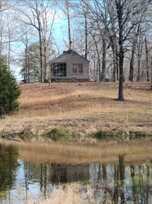 A small wooden cabin on a hill, surrounded by bare trees, reflecting in a calm pond below.