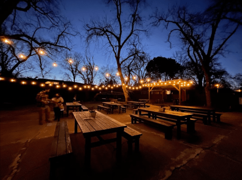 A dimly lit outdoor space with wooden tables, string lights, and bare trees at dusk. Two people are walking in the background.