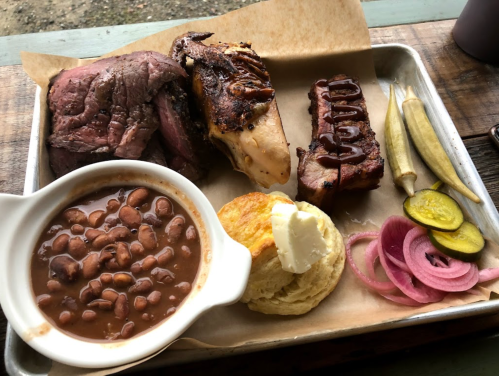 A tray with smoked meat, baked beans, a biscuit with butter, pickles, and pickled onions.