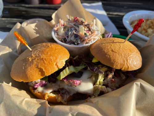 Two sandwiches with melted cheese and coleslaw on a plate, accompanied by a small bowl of coleslaw.