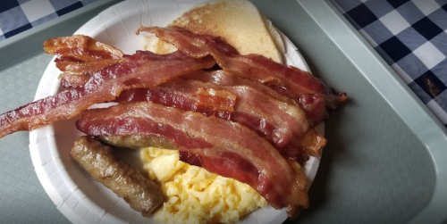 A plate with crispy bacon strips, scrambled eggs, and a sausage link, served on a paper plate.