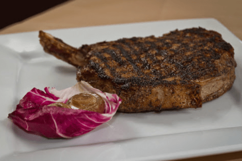 A grilled ribeye steak with a charred crust, served on a white plate alongside a slice of radicchio.