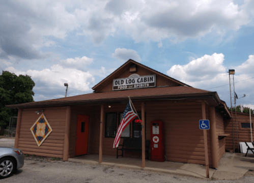 A rustic log cabin restaurant with an American flag, "OPEN" sign, and a vintage gas pump outside under a cloudy sky.