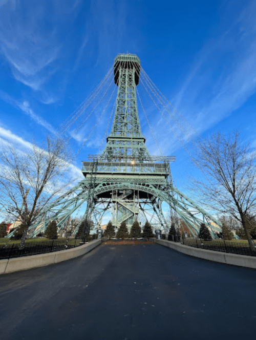 A view of a green metal tower resembling the Eiffel Tower against a blue sky with wispy clouds and bare trees.