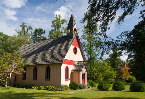 A quaint church with a steeple, surrounded by lush greenery and trees under a blue sky with scattered clouds.