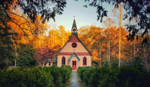 A quaint church surrounded by trees with autumn foliage, featuring a pointed steeple and a path leading to the entrance.