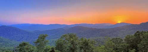 A panoramic view of mountains at sunset, with vibrant colors illuminating the sky and lush greenery in the foreground.