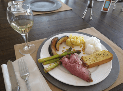 A plate with roast beef, asparagus, squash, mashed potatoes, and garlic bread, accompanied by a glass of white wine.